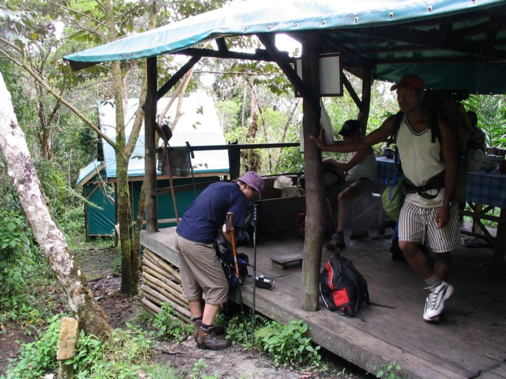 Several people are loading their backpacks at a wooden shelter in the rainforest. A green canvas-covered cabin is in the background.