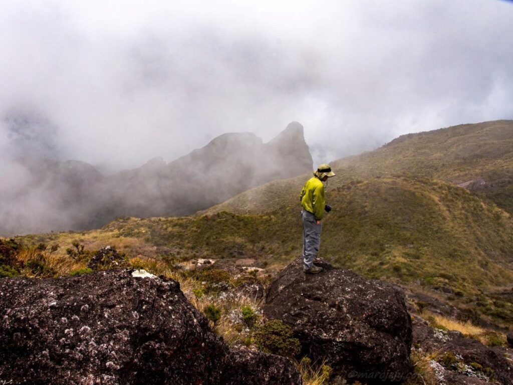 A man in a lime-green jacket stands on a boulder looking out over mist-shrouded peaks and hills