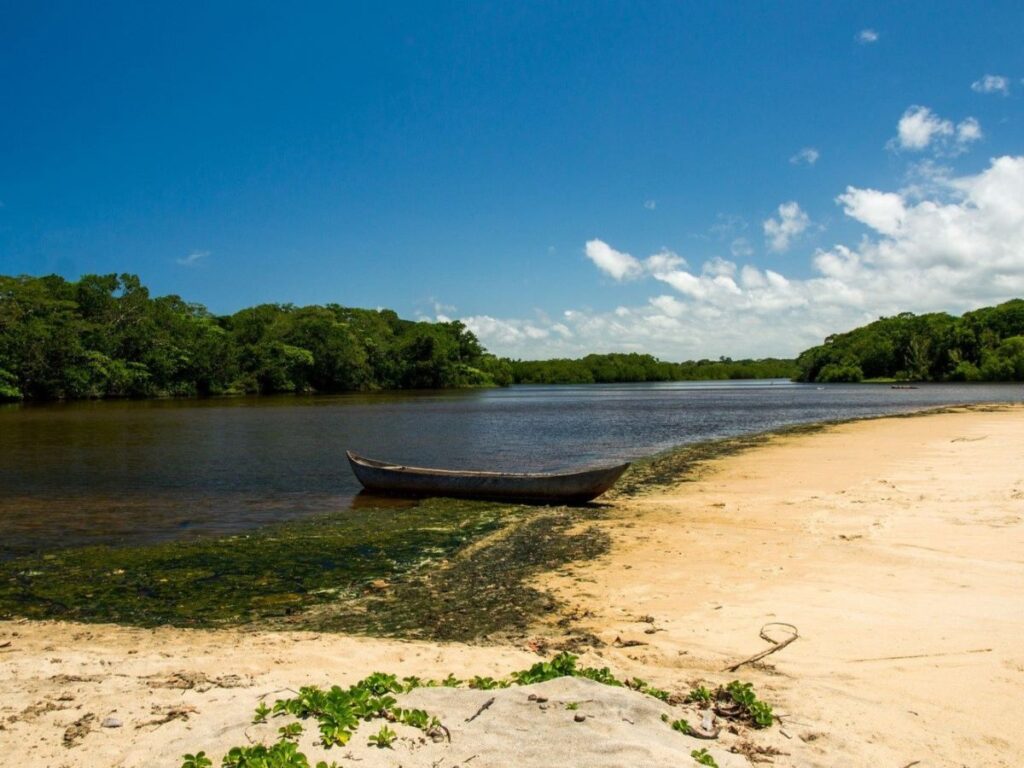 Une pirogue est tirée jusqu'à la rive sablonneuse d'une rivière large et calme. Les bords de la rivière sont bordés d'une végétation dense.