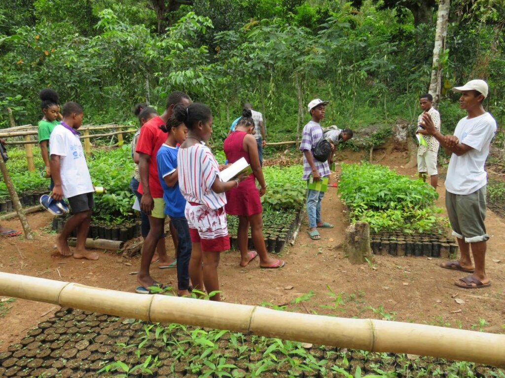 Un homme parle à 11 jeunes étudiants dans une pépinière. La pépinière est située dans une clairière de la forêt. Elle contient des centaines de petits pots contenant des semis d'arbres.