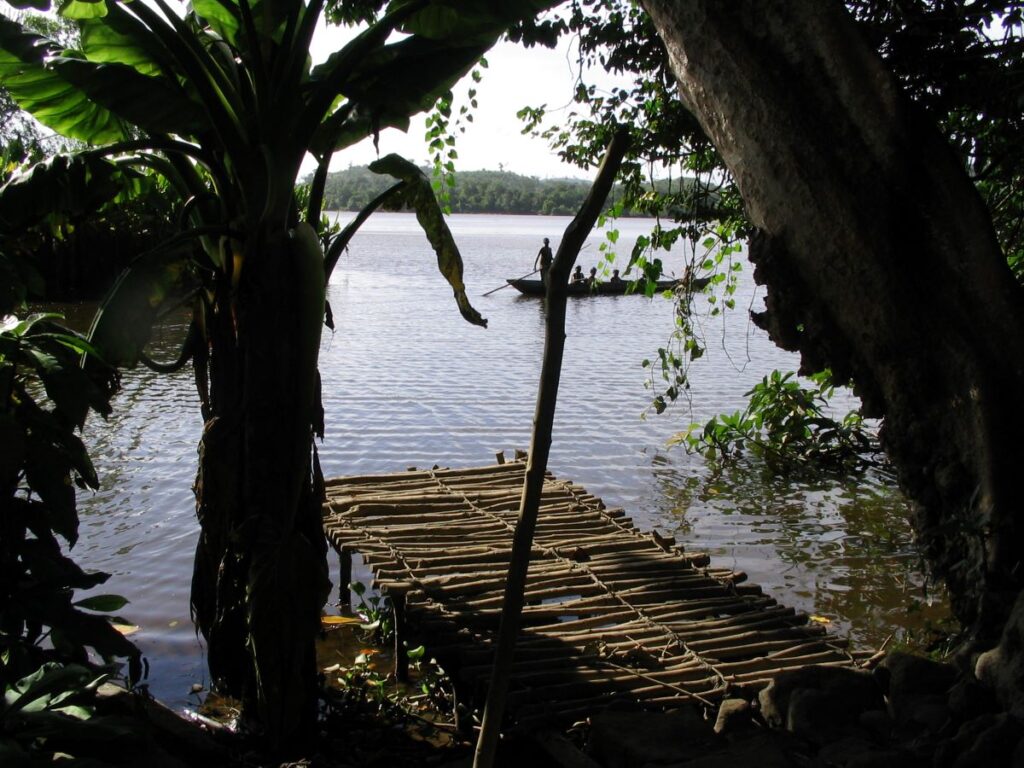 Un quai à bâtons rompus s'étend sur quelques mètres dans une rivière depuis les rives ombragées et bordées d'arbres ; la rivière est très large et une pirogue avec cinq personnes à bord s'approche du quai.