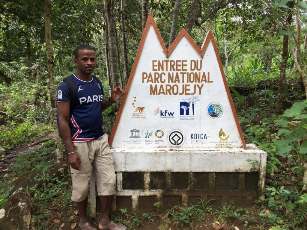 A park guide stands next to a big sign in the forest that says "Entrée du Parc National Marojejy".