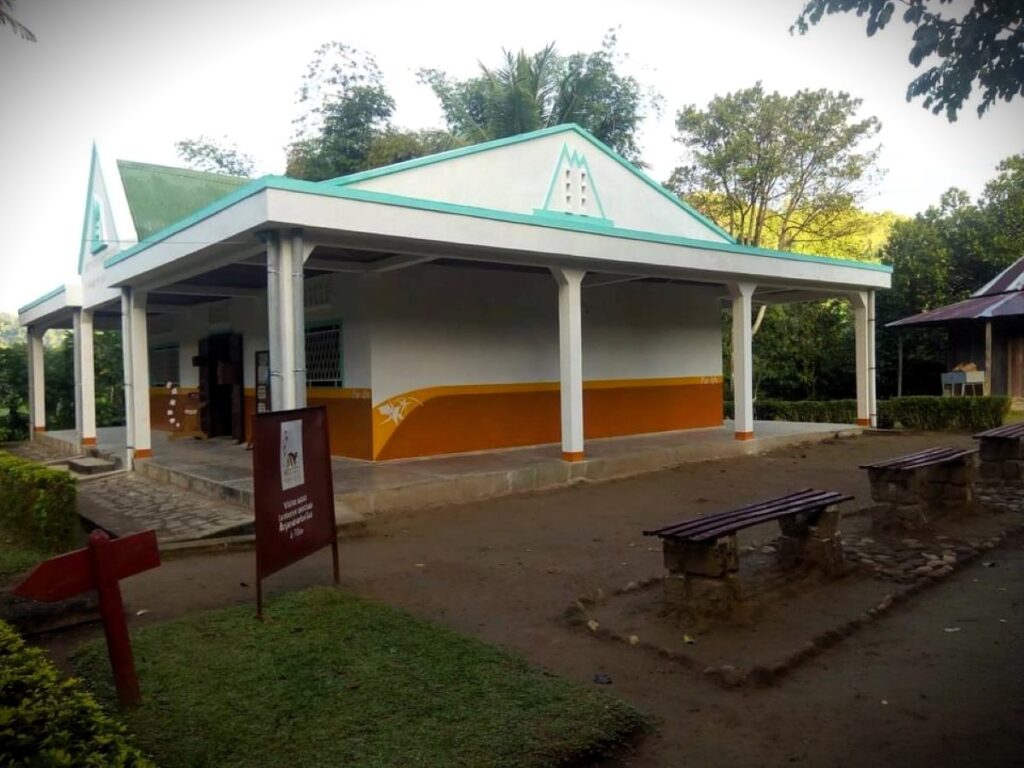 A white building with brown and turquoise trim and a wide veranda. There are benches and a few signs on the side of the building.