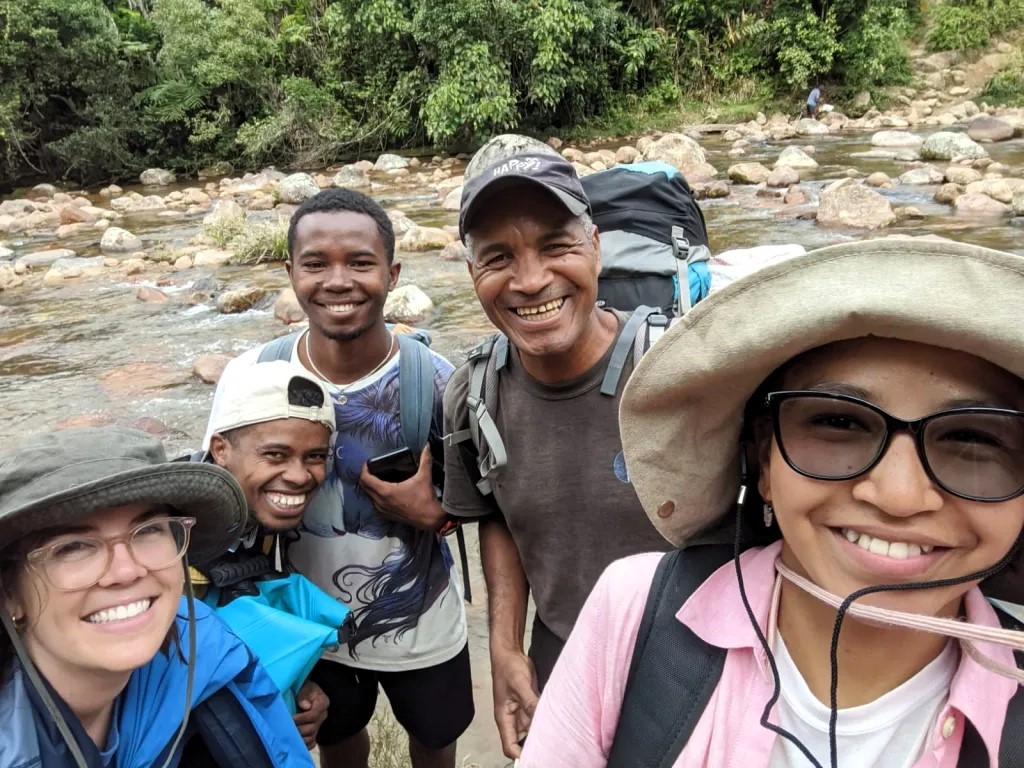 Five people are smiling at the camera. A rocky riverbed is in the background.