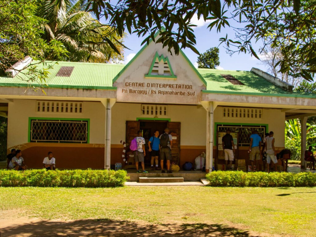 Numerous people stand on the front porch of a building with a pitched green roof and a sign that reads "Centre d'Interpretation, PN Marojejy / RS Anjanaharibe-Sud".