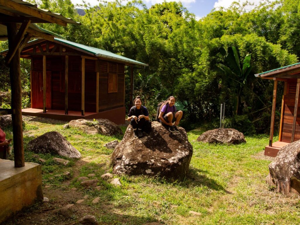 Photo de deux personnes assises sur un gros rocher, entre de petites cabanes, au cœur d'une forêt dense de bambous.